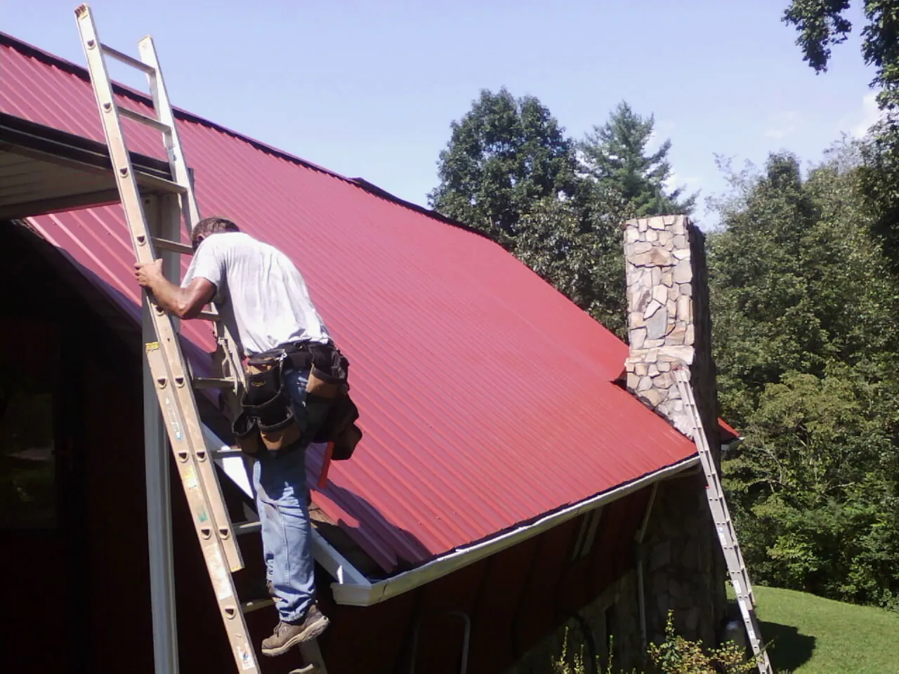 Roofer on a ladder beside a red metal roof and stone chimney — Soffit & Fascia Repair by Roofing Brothers in Riverhead